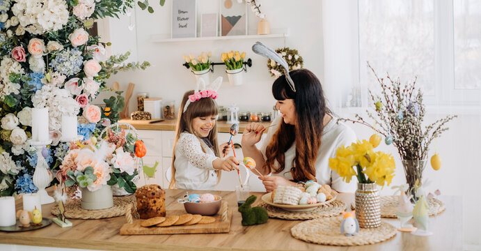 Cute little girl in bunny ears with her mother paint eggs while sitting at the table in the kitchen. Decorations for the celebration of Easter