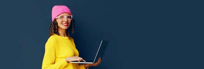 Naklejka premium Portrait of stylish modern young woman working with laptop wearing colorful clothes on dark blue background, banner blank copy space for advertising text