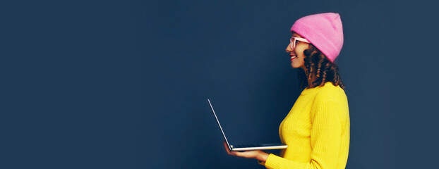 Portrait of stylish modern young woman working with laptop wearing colorful clothes on dark blue background, banner blank copy space for advertising text