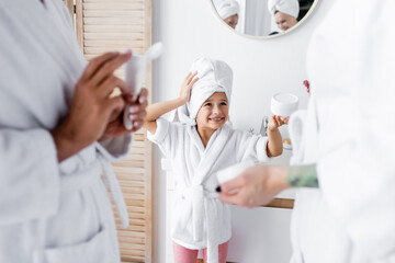 Positive kid in towel and bathrobe holding cosmetic cream near mothers in bathroom