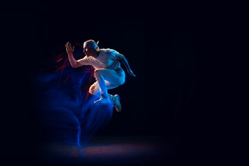 Fototapeta premium Jumping. Stylish man in sports white outfit dancing hip-hop, breakdance isolated on dark background in mixed neon light. Youth culture, hip-hop, style and fashion, action.