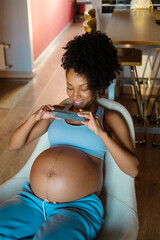 Cheerful black pregnant woman recording video or taking photos of her belly at home.
