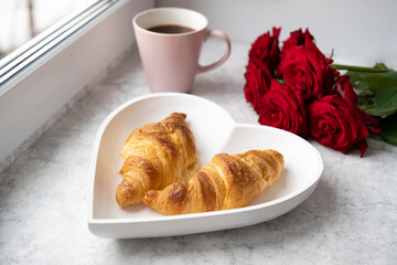 Beautiful romantic breakfast.French croissants on white heart-shaped plate , roses and cup of coffee on windowsill. Close-up.Valentine day,love or dating concept