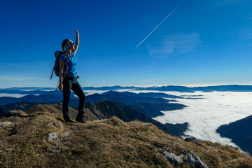 Obraz premium Woman with backpack hiking and admiring a scenic view from mount Eisenerzer Reichenstein in Styria, Austria, Europe. Ennstal valley is covered in clouds. Hiking trail, Wanderlust. Freedom concept
