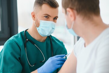 medicine, vaccination and healthcare concept - doctor wearing face protective medical mask for protection from virus disease with syringe doing injection of vaccine to male patient