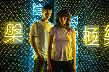 Young couple in white t-shirts stand against the background of a metal grid with neon symbols.