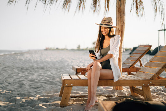 A young girl in a tropical resort during her vacation sits on a wooden deck chair near straw umbrellas on the beach, holds a smartphone in her hands and makes a video call using the application.