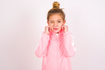 caucasian little kid girl with bun hairstyle wearing pink tracksuit over white background  covering ears with fingers with annoyed expression for the noise of loud music. Deaf concept.