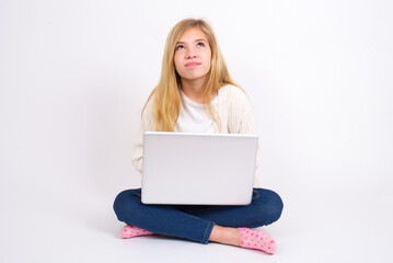 caucasian teen girl sitting with laptop in lotus position on white background has worried face looking up lips together, being upset thinking about something important, keeps hands down.