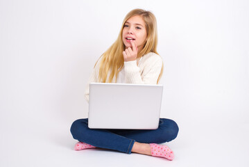 Fototapeta premium caucasian teen girl sitting with laptop in lotus position on white background with thoughtful expression, looks to the camera, keeps hand near face, bitting a finger thinks about something pleasant.