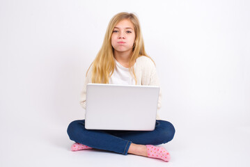 caucasian teen girl sitting with laptop in lotus position on white background puffing cheeks with funny face. Mouth inflated with air, crazy expression. - obrazy, fototapety, plakaty