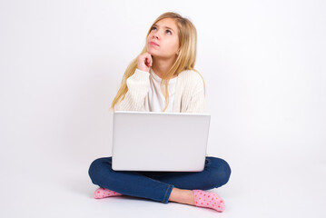 Portrait of thoughtful caucasian teen girl sitting with laptop in lotus position on white background keeps hand under chin, looks away trying to remember something or listens something with interest.