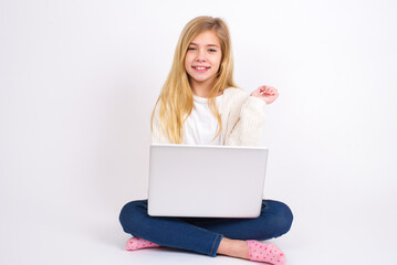 Cheerful caucasian teen girl sitting with laptop in lotus position on white background with hand near face. Looking with glad expression at the camera after listening to good news. Confidence.