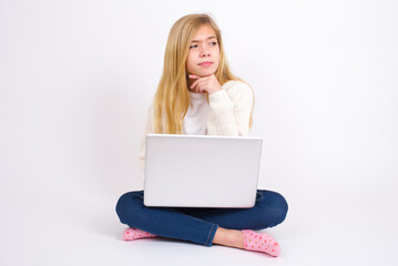 Dreamy caucasian teen girl sitting with laptop in lotus position on white background with pleasant expression, looks sideways, keeps hand under chin, thinks about something pleasant.