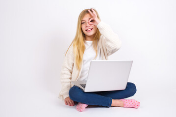 caucasian teen girl sitting with laptop in lotus position on white background with happy face smiling doing ok sign with hand on eye looking through finger.