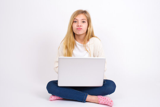 caucasian teen girl sitting with laptop in lotus position on white background crosses eyes, puts lips, makes grimace with awkward expression has fun alone, plays fool.