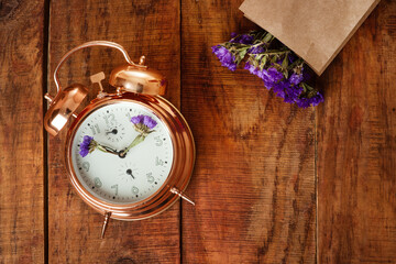 gold alarm clock with blue flowers instead of clock's hands and paper bag with flowers on wooding surface. Spring time
