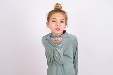 little caucasian kid girl with hair bun wearing technical shirt over white background looking at the camera blowing a kiss with hand on air being lovely and sexy. Love expression.