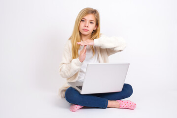 caucasian teen girl sitting with laptop in lotus position on white background feels tired and bored, making a timeout gesture, needs to stop because of work stress, time concept.