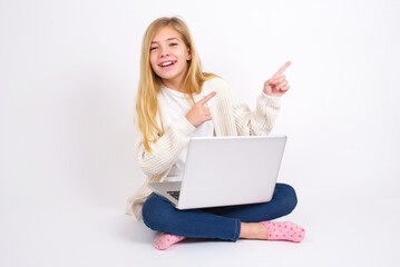 caucasian teen girl sitting with laptop in lotus position on white background with positive expression, indicates with fore finger at blank copy space for your promotional text or advertisement.
