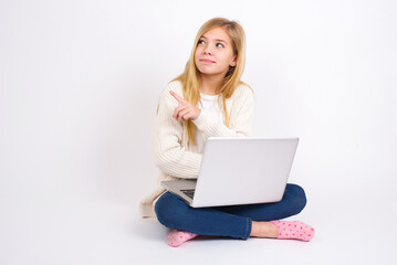 Fototapeta premium Portrait of caucasian teen girl sitting with laptop in lotus position on white background posing on camera with tricky look, presenting product with index finger. Advertisement concept.