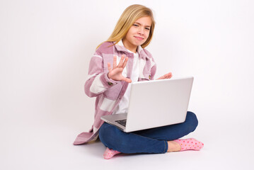 Naklejka premium Afraid caucasian teen girl sitting with laptop in lotus position on white background , makes terrified expression and stop gesture with both hands saying: Stay there. Panic concept.