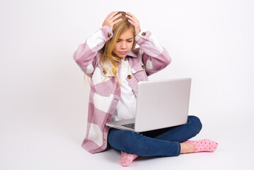 caucasian teen girl sitting with laptop in lotus position on white background holding head with hands, suffering from severe headache, pressing fingers to temples