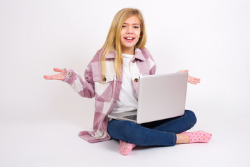 Naklejka premium So what? Portrait of arrogant caucasian teen girl sitting with laptop in lotus position on white background shrugging hands sideways smiling gasping indifferent, telling something obvious.