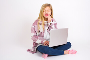 Fototapeta premium Tooth ache concept. caucasian teen girl sitting with laptop in lotus position feeling pain, holding his cheek with hand, suffering from bad toothache, looking at camera with painful expression