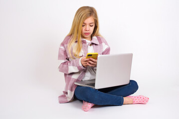 Smiling  caucasian teen girl sitting with laptop in lotus position on white using cell phone, messaging, being happy to text with friends, looking at smartphone. Modern technologies and communication.