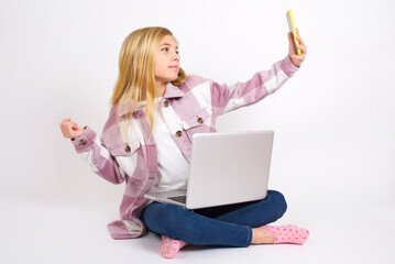 Portrait of a  caucasian teen girl sitting with laptop in lotus position on white background taking a selfie to send it to friends and followers or post it on his social media.