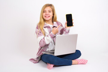 Smiling  caucasian teen girl sitting with laptop in lotus position on white background showing and pointing at empty phone screen. Advertisement and communication concept.