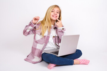  caucasian teen girl sitting with laptop in lotus position on white background holding an invisible aligner and pointing to her perfect straight teeth. Dental healthcare and confidence concept.