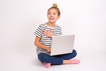 Joyful caucasian teen girl sitting with laptop in lotus position on white background  expresses positive emotions recalls something funny keeps hand on chest and giggles happily.