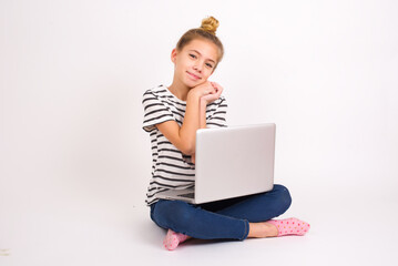 Charming serious caucasian teen girl sitting with laptop in lotus position on white background keeps hands near face smiles tenderly at camera