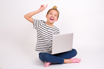 Photo of upbeat caucasian teen girl sitting with laptop in lotus position on white background has fun and dances carefree wear being in perfect mood makes movements. Spends free time on disco party