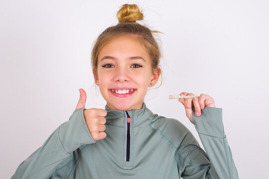 little caucasian kid girl with hair bun wearing technical shirt over white wall holding an invisible braces aligner and rising thumb up, recommending this new treatment. Dental healthcare concept