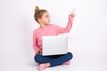 caucasian teen girl sitting with laptop in lotus position on white background pointing to object on copy space, rear view. Turn your back