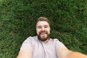 young man with beard and fashion hair style lying on grass taking selfie - holding smart phone or tablet and looking at camera.