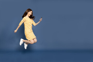Portrait of Asian woman jumping and using mobile phone application isolated over blue background,...