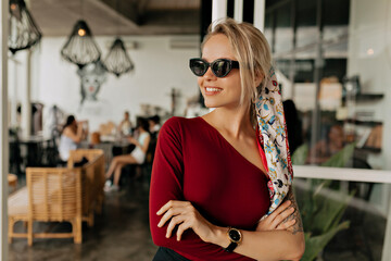 Obraz na płótnie Canvas Attractive pretty woman in sunglasses wearing bare shoulder sweater, sunglasses and head accessory is looking away and smiling on background of summer modern cafeteria