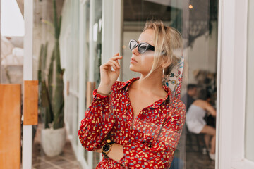 Attractive stylish young woman in sunglasses and bright summer dress is posing under glass door while resting in summer modern cafeteria. Pretty woman is having breakfast outdoor