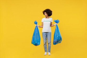 Full body young woman of African American ethnicity in white volunteer t-shirt hold trash bags after rubbish removing isolated on plain yellow background. Voluntary free work assistance help concept