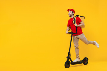 Full body side view delivery guy employee man in red cap T-shirt uniform workwear work as dealer courier ride electric kick scooter hold thermal food bag backpack isolated on plain yellow background.