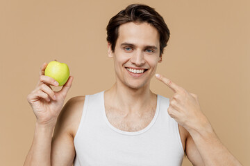 Attractive fun young man 20s perfect skin in undershirt hold apple point finger on white teeth smile isolated on pastel pastel beige background studio Skin care healthcare cosmetic procedures concept