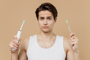 Attractive puzzled pensive young man 20s perfect skin wear undershirt hold choice electric brushes...
