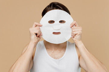 Attractive young man 20s perfect skin in undershirt hold cover face with tissue fabric textile face mask isolated on pastel pastel beige background. Skin care healthcare cosmetic procedures concept.