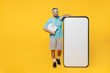 Full body fun young fitness trainer instructor sporty man sportsman in blue t-shirt big screen mobile cell phone with workspace hold scales isolated on plain yellow background. Workout sport concept.