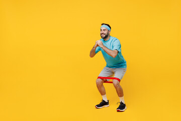 Naklejka na ściany i meble Full body side view happy young fitness trainer instructor sporty man sportsman in headband blue t-shirt use fitness elastic bands do squats isolated on plain yellow background. Workout sport concept.