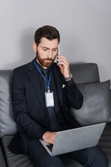 bearded businessman with id badge talking on smartphone and using laptop in hotel room